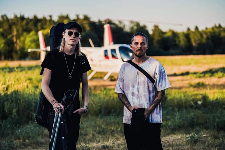 Två artister står på en äng framför en helikopter